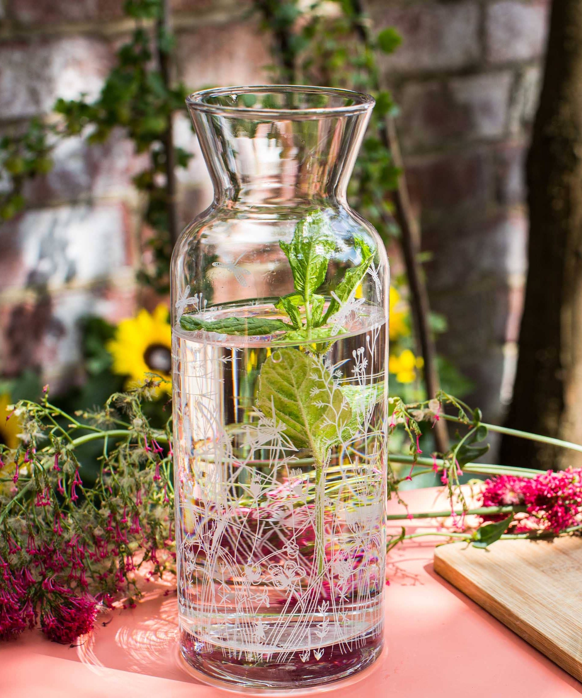 Glass carafe with water and herbs on a table with flowers and a cutting board in the background.