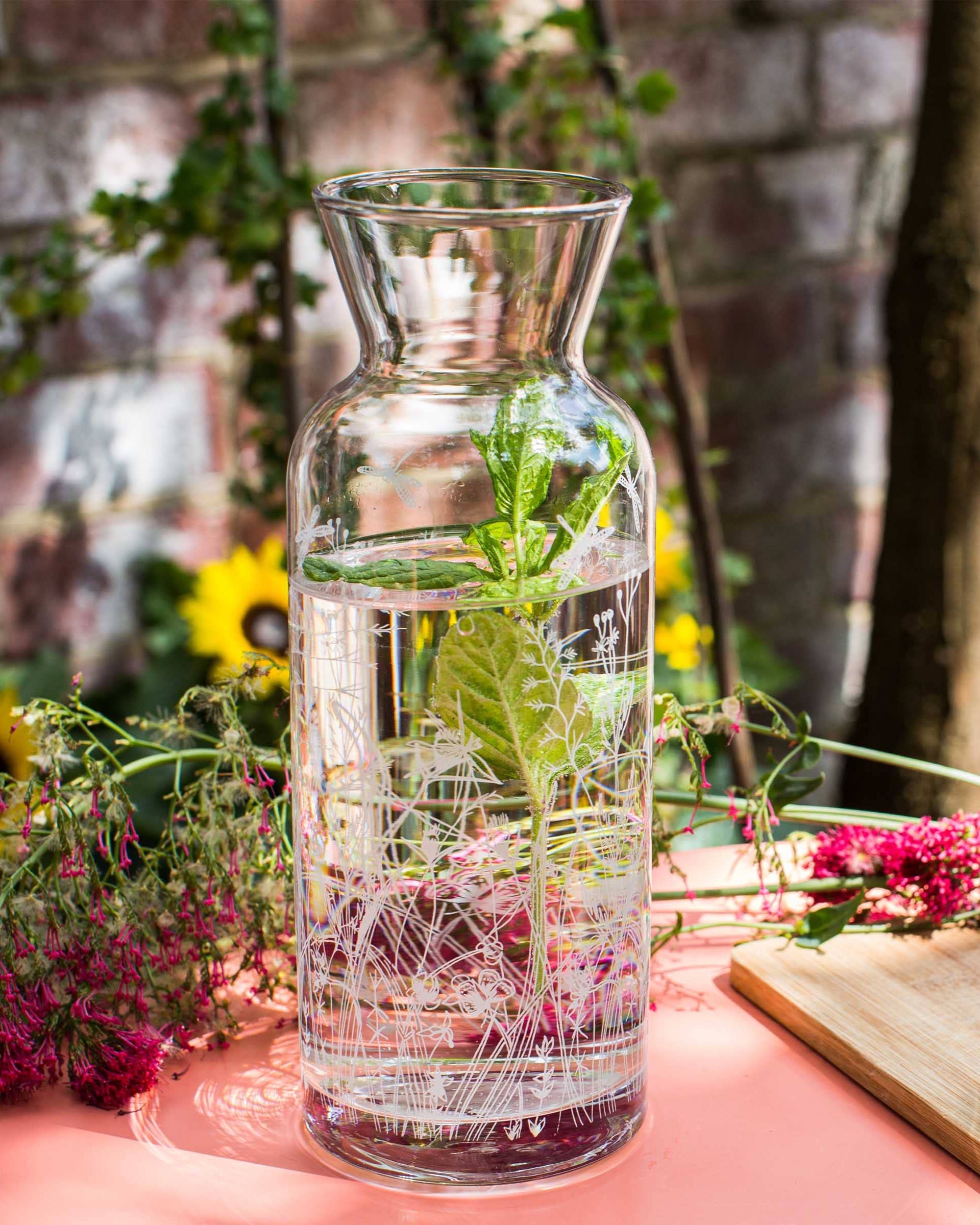Glass carafe with water and herbs on a table with flowers and a cutting board in the background.