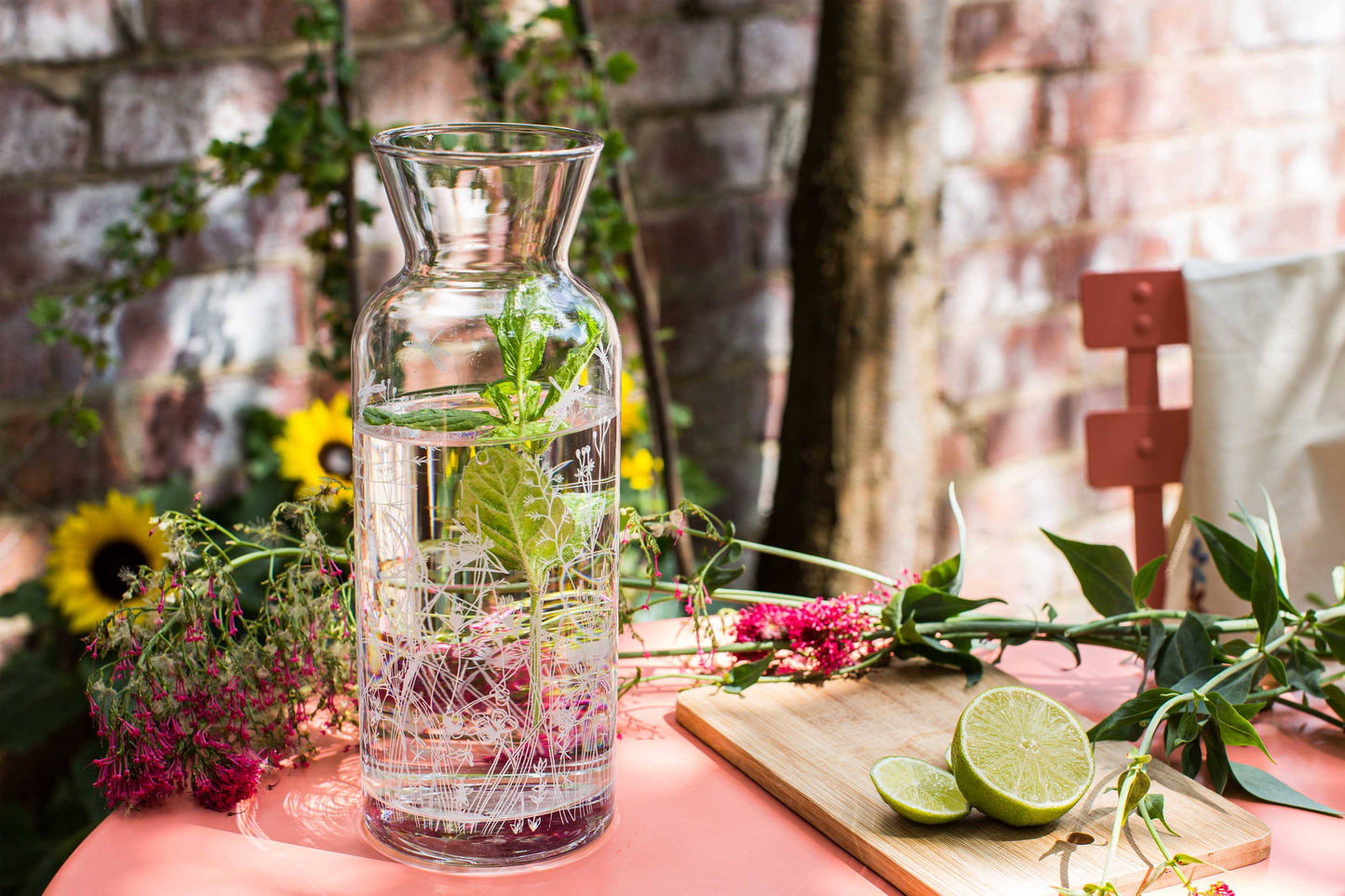 Glass carafe with water and herbs on a table with flowers and a cutting board in the background.
