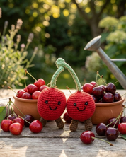 Two red crocheted cherry characters with green stems and smiling faces stand in front of terracotta bowls filled with real cherries on a wooden table, with a metal watering can and garden plants blurred in the background.