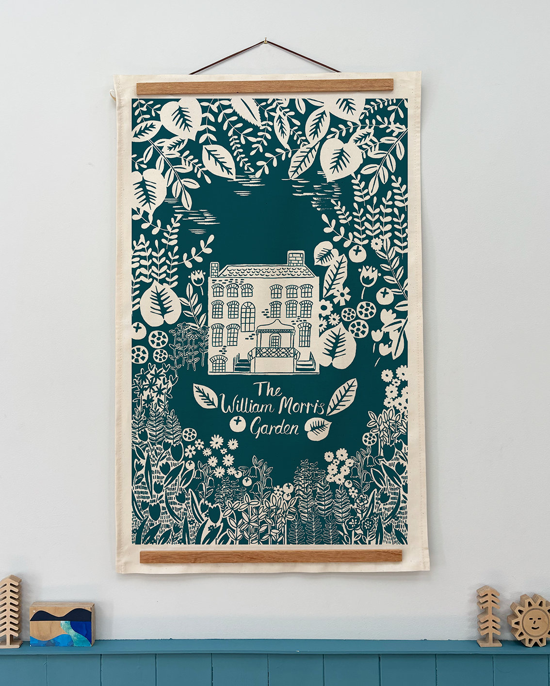Decorative tea towel with a house design and floral elements on a teal background, framed and hanging on a wall.