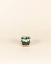 Small ceramic cup with green and blue glaze on a beige background