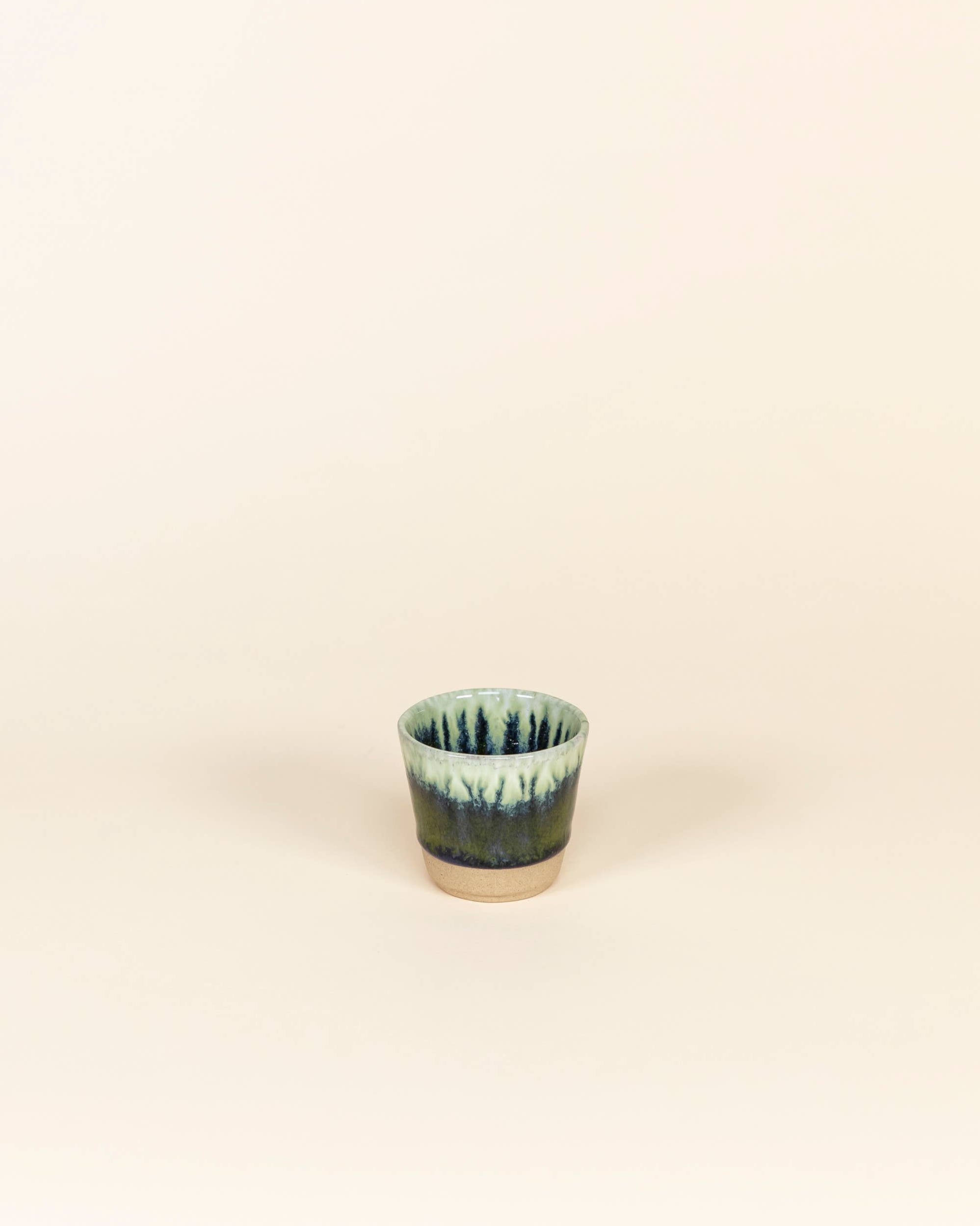 Small ceramic cup with green and blue glaze on a beige background