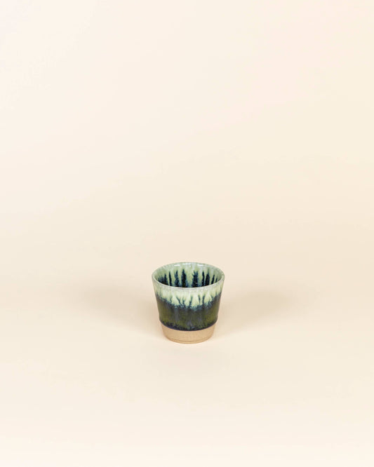 Small ceramic cup with green and blue glaze on a beige background