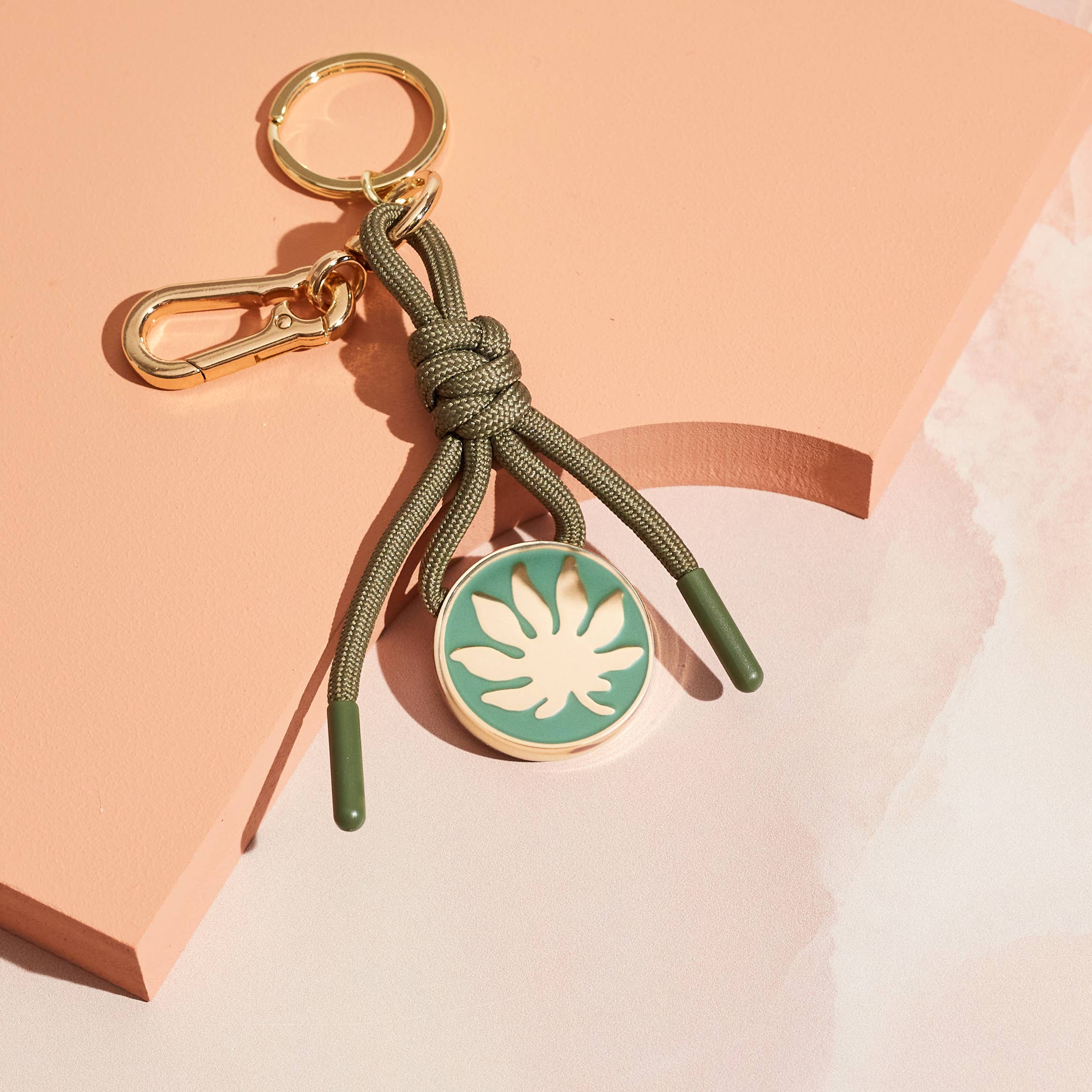 Bag charm keychain with green rope and circular leaf charm in gold on green.