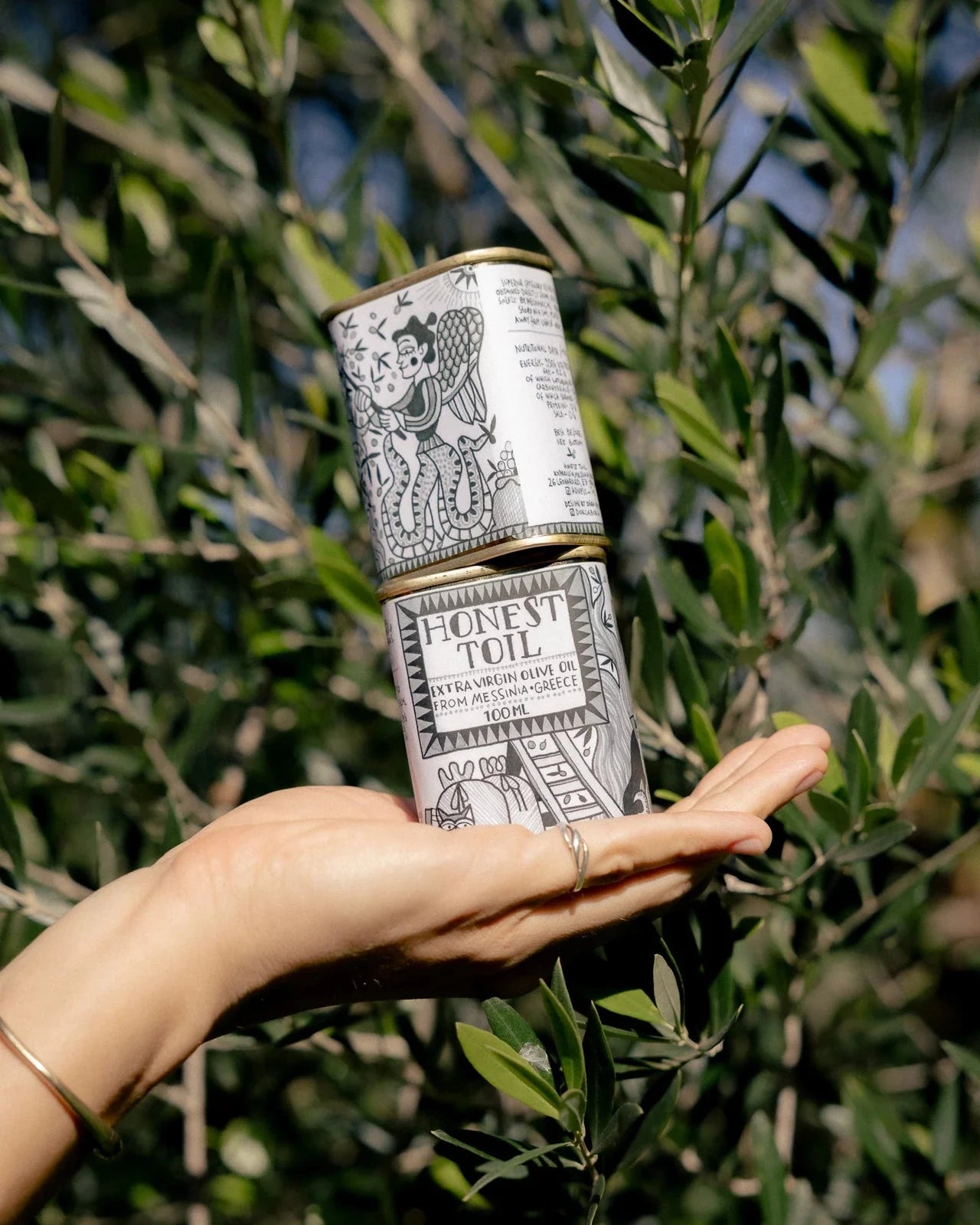 A hand holding a stack of two cylindrical metal tins of Honey Toile Extra Virgin Rose Hip and Black Seed Oils in front of a leafy green plant.