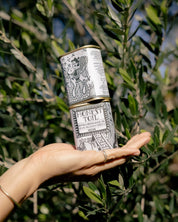 A hand holding a stack of two cylindrical metal tins of Honey Toile Extra Virgin Rose Hip and Black Seed Oils in front of a leafy green plant.
