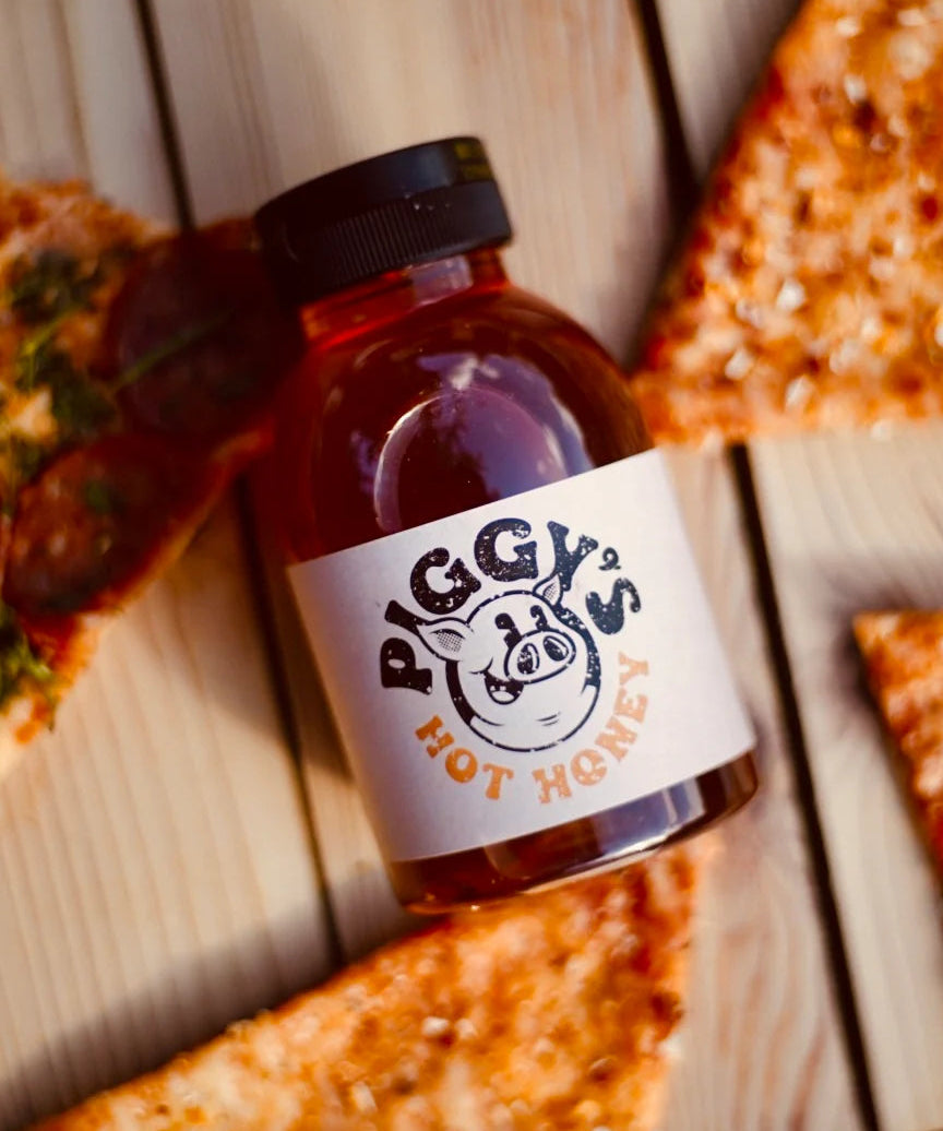 Bottle of 'Piggy's Hot Honey' on a wooden surface with pizza slices.