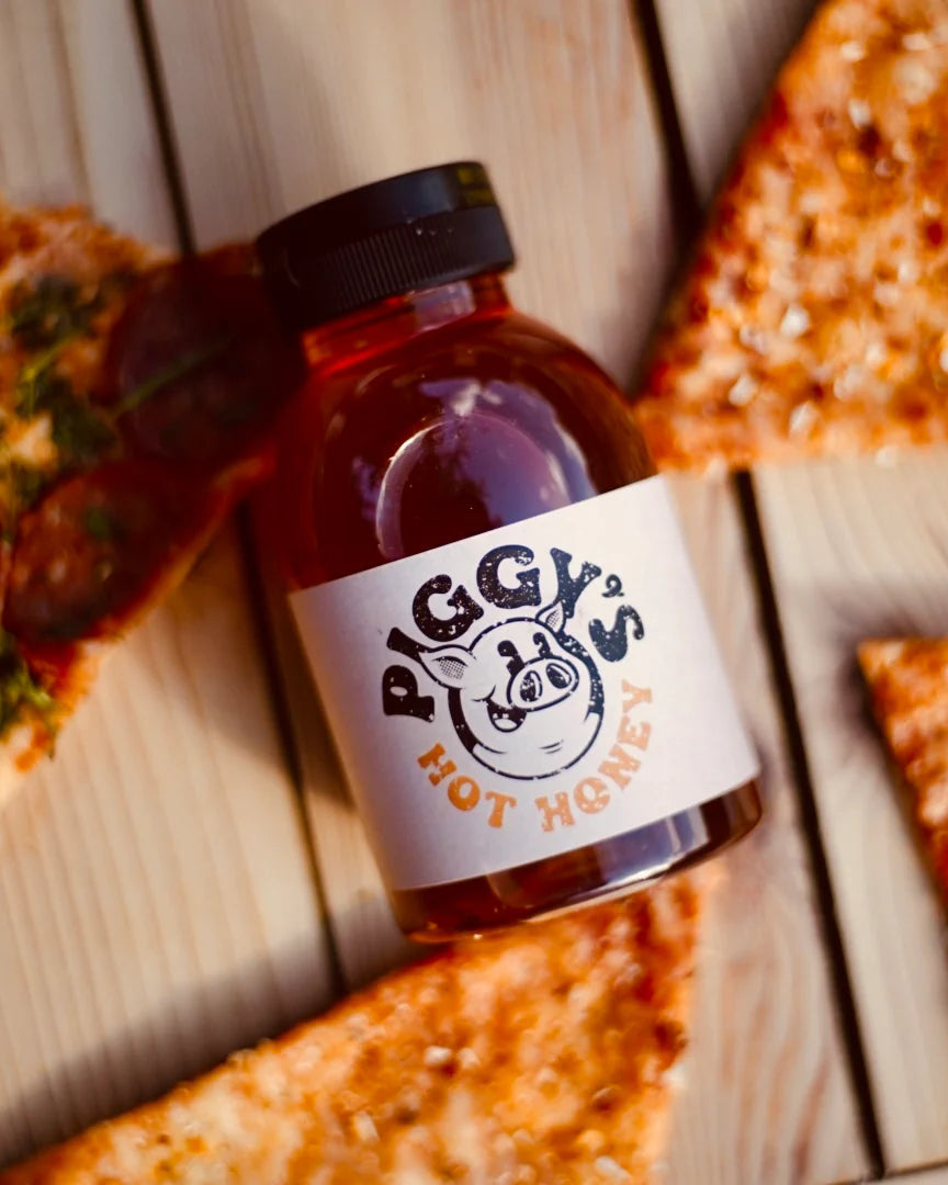 Bottle of 'Piggy's Hot Honey' on a wooden surface with pizza slices.